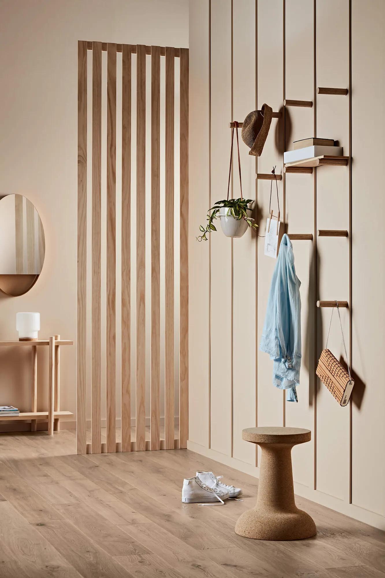 A modern interior space featuring an open wooden wall divider with horizontal slats, a hanging white pendant lamp, a wooden bench with personal items, shelves mounted on the wall divider holding books and decorative items, a potted plant, and a blue shirt hanging neatly over the shelf. The floor appears to be light-colored hardwood or laminate, and there is a soft focus on the room's simplicity and tranquility