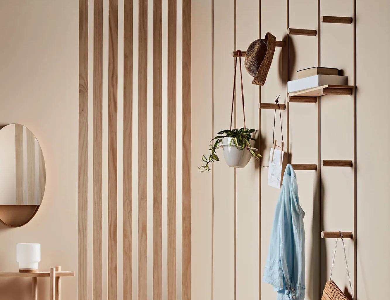 A modern interior space featuring an open wooden wall divider with horizontal slats, a hanging white pendant lamp, a wooden bench with personal items, shelves mounted on the wall divider holding books and decorative items, a potted plant, and a blue shirt hanging neatly over the shelf. The floor appears to be light-colored hardwood or laminate, and there is a soft focus on the room's simplicity and tranquility
