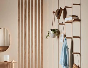 A modern interior space featuring an open wooden wall divider with horizontal slats, a hanging white pendant lamp, a wooden bench with personal items, shelves mounted on the wall divider holding books and decorative items, a potted plant, and a blue shirt hanging neatly over the shelf. The floor appears to be light-colored hardwood or laminate, and there is a soft focus on the room's simplicity and tranquility