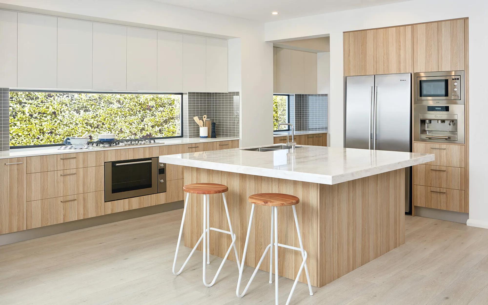 A spacious kitchen with light wooden cabinets, white countertops, a steel refrigerator, stainless steel appliances, and modern furniture
