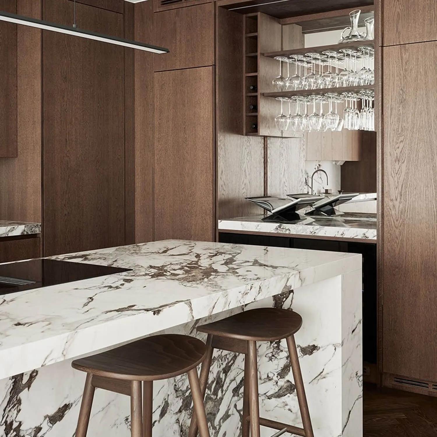 A well-lit kitchen interior featuring a modern design, marble countertops, wood cabinets, a wine glass rack, and stainless steel appliances
