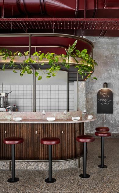 Restaurant interior with red stools, a wooden bar, hanging lights, and wall shelving