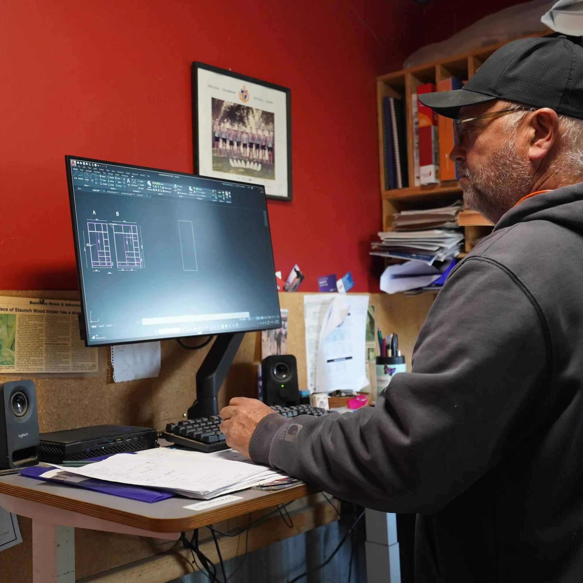 A person works at a desk using CAD software in a home office with shelving, documents and computer equipment.