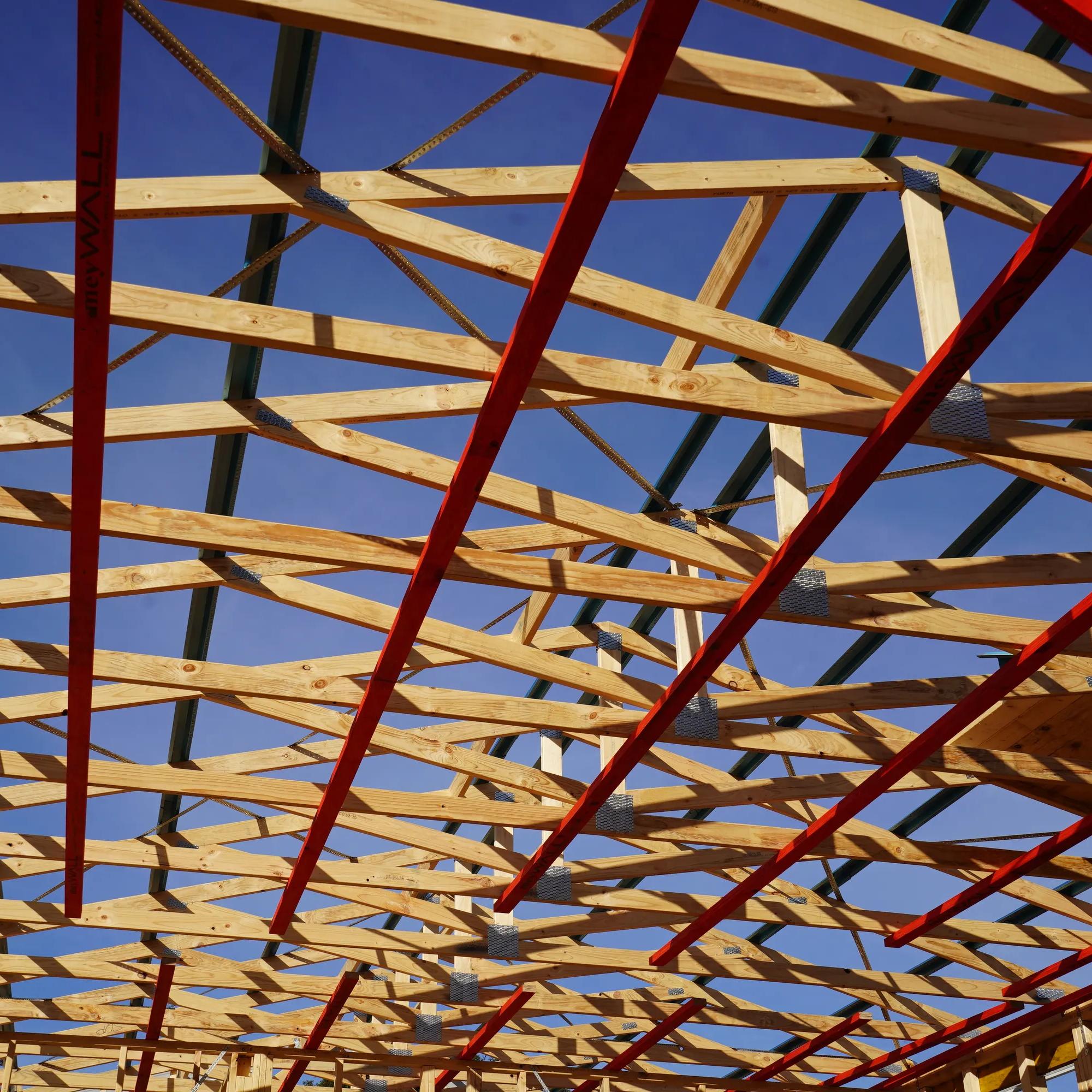 Timber roof trusses and red structural braces viewed from below against a clear blue sky
