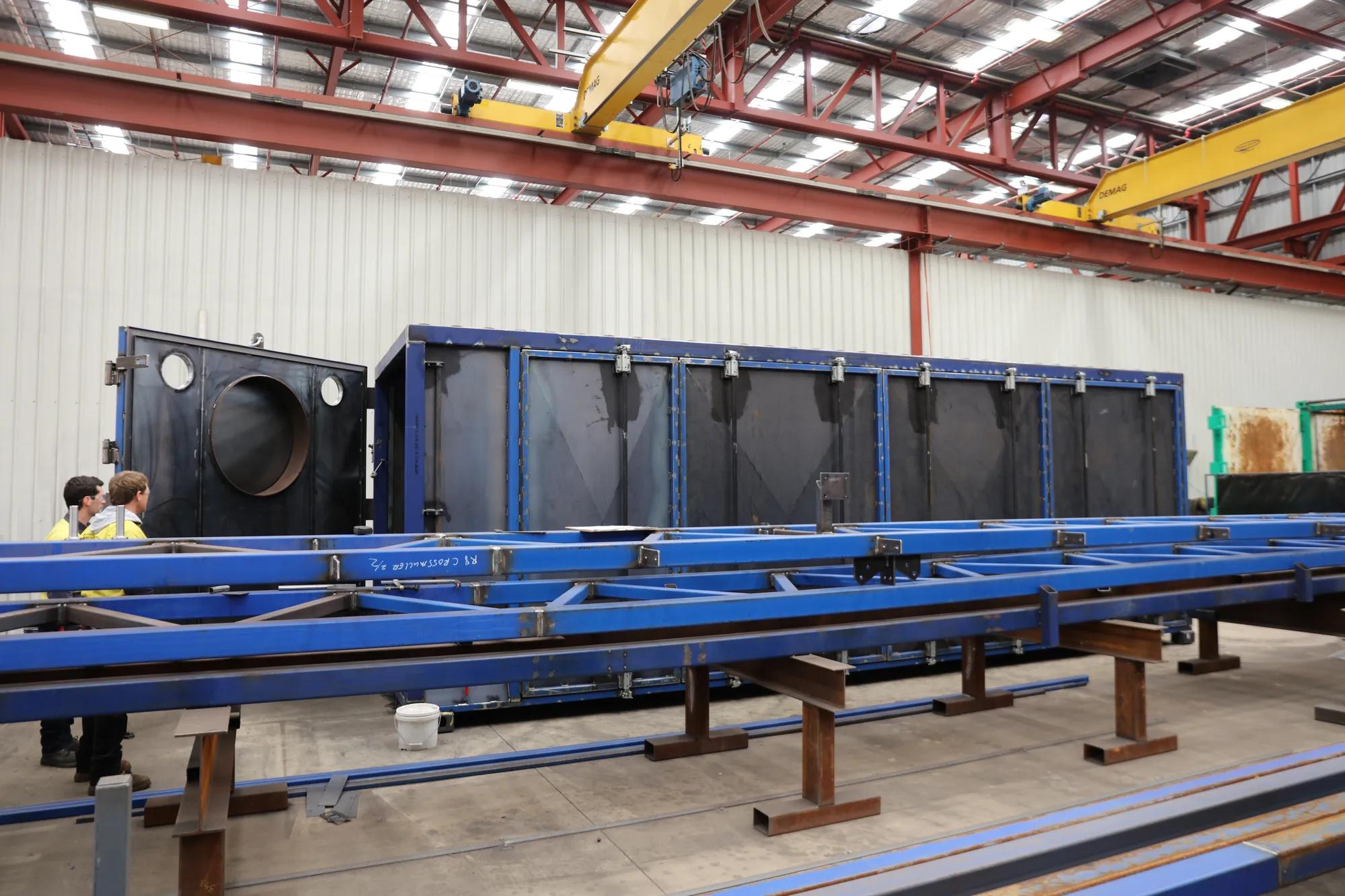 Large blue steel fabricated structure inside industrial warehouse with cranes.