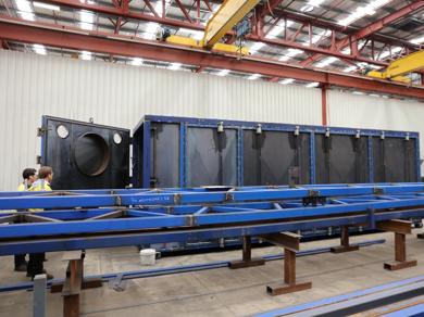 Large blue steel fabricated structure inside industrial warehouse with cranes.