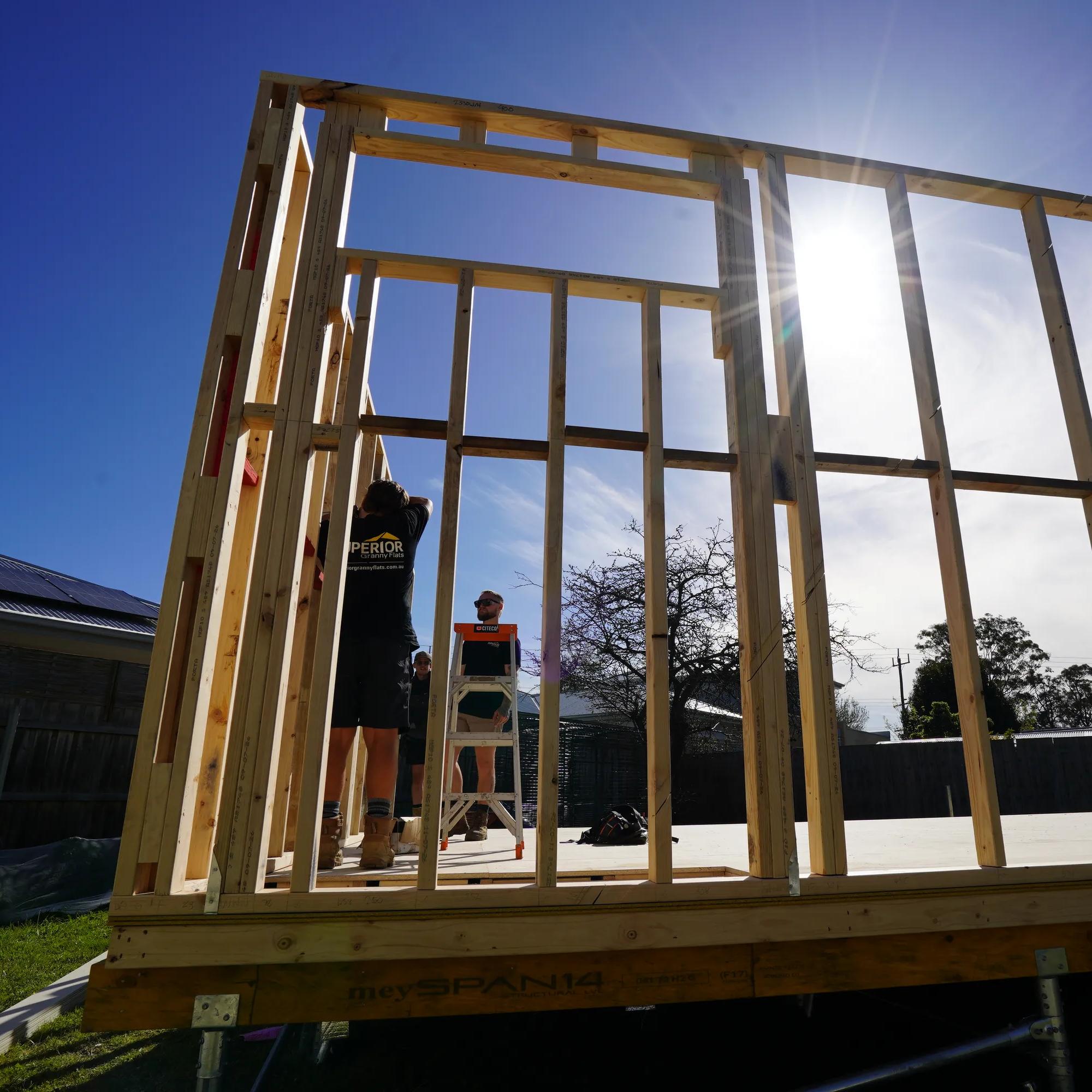 Timber wall framing under construction on a raised outdoor platform with workers