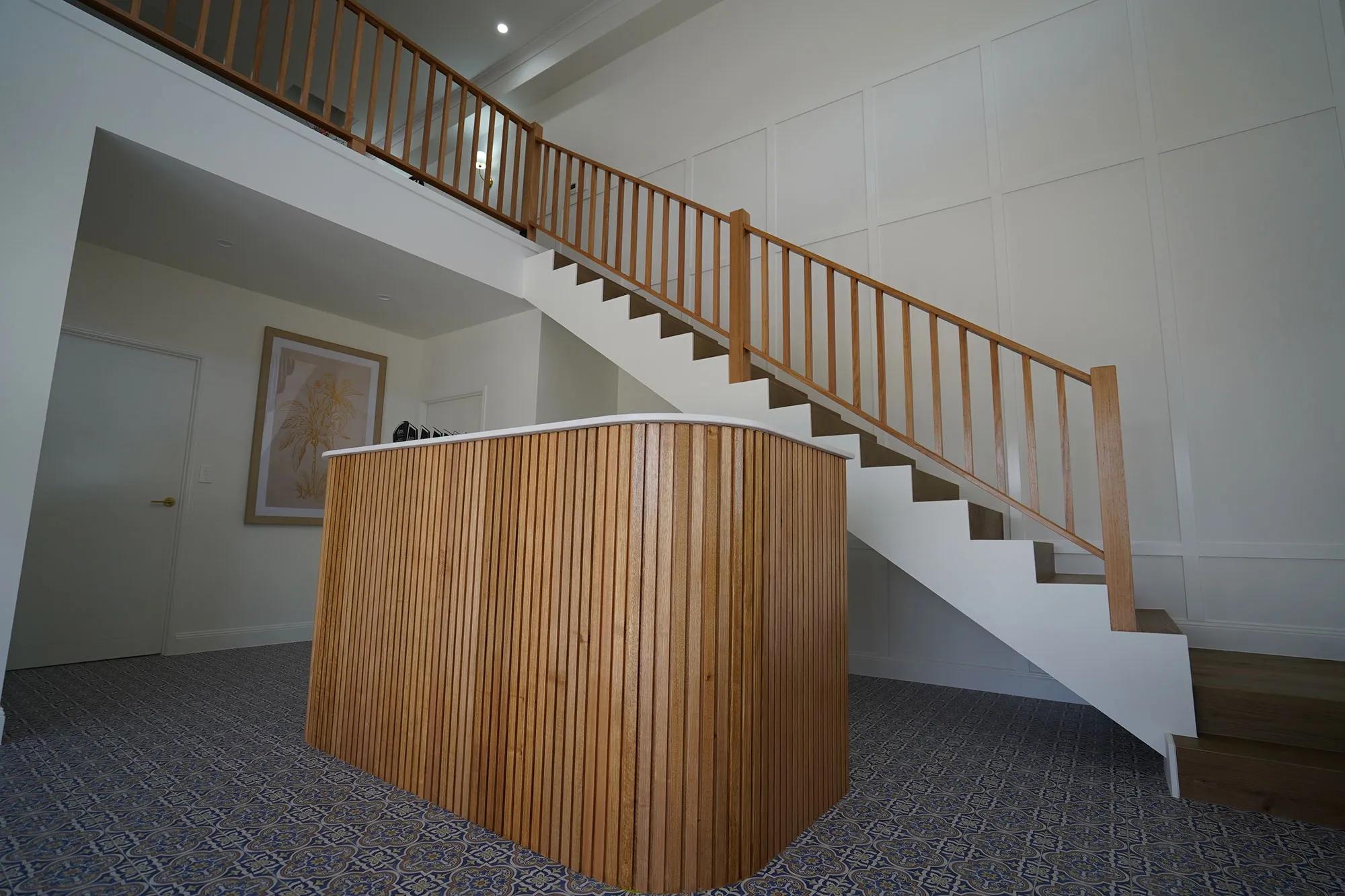 An interior space, specifically a stairway. The staircase features light wood banisters on both sides. At the top, there's a modern-looking room that includes a desk or counter area, which appears to be part of a reception area given its placement and design. Below the stairs, the floor is carpeted with a neutral color. There are no visible texts in this image