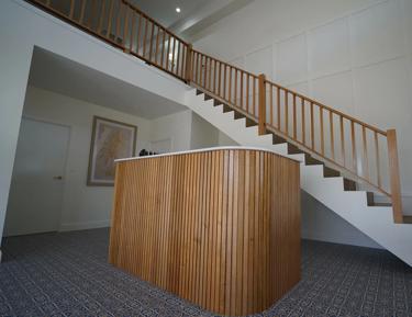 An interior space, specifically a stairway. The staircase features light wood banisters on both sides. At the top, there's a modern-looking room that includes a desk or counter area, which appears to be part of a reception area given its placement and design. Below the stairs, the floor is carpeted with a neutral color. There are no visible texts in this image