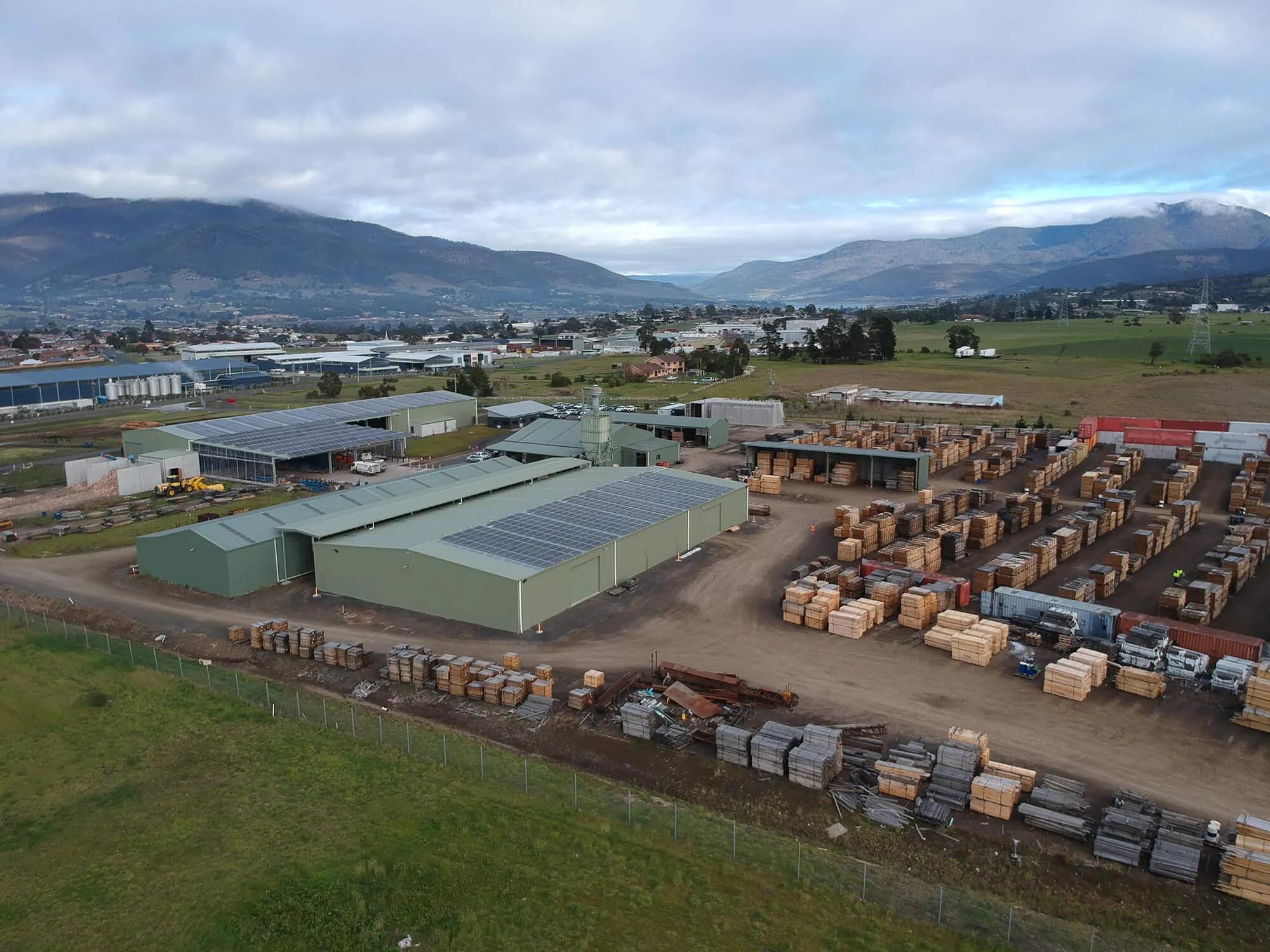 This is a picture of a small industrial area in a rural setting. The foreground is dominated by a large metal shed filled with stacks of wooden pallets. Behind this, there's another building with what appears to be some kind of material storage or production taking place. In the background, you can see a few other buildings and a truck parked outside. There's also a mountain range visible in the distance under a partly cloudy sky. The overall scene suggests an outdoor industrial or warehouse area