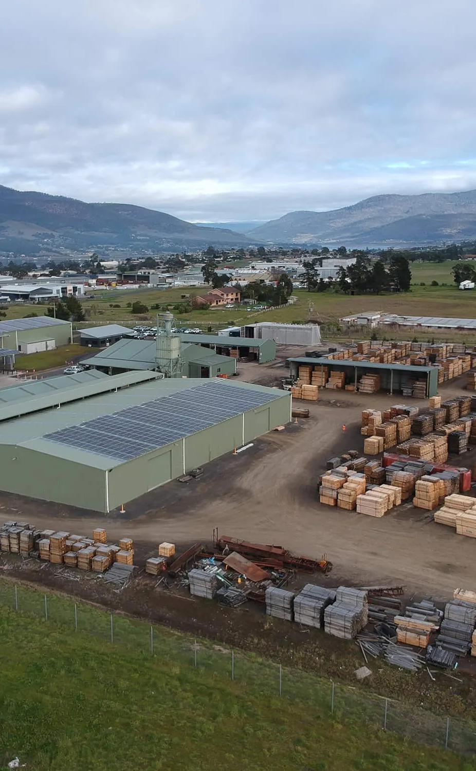 This is a picture of a small industrial area in a rural setting. The foreground is dominated by a large metal shed filled with stacks of wooden pallets. Behind this, there's another building with what appears to be some kind of material storage or production taking place. In the background, you can see a few other buildings and a truck parked outside. There's also a mountain range visible in the distance under a partly cloudy sky. The overall scene suggests an outdoor industrial or warehouse area