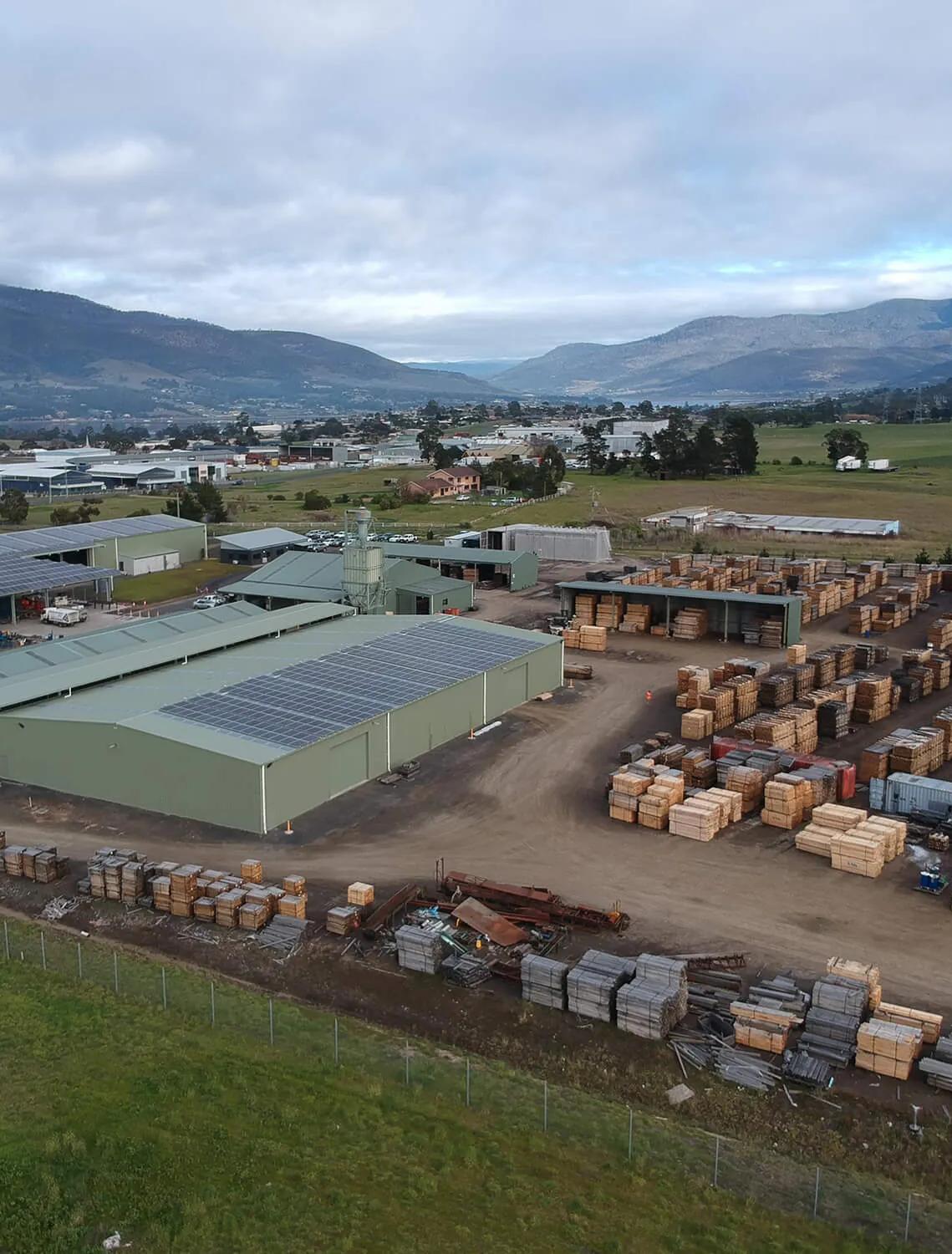 This is a picture of a small industrial area in a rural setting. The foreground is dominated by a large metal shed filled with stacks of wooden pallets. Behind this, there's another building with what appears to be some kind of material storage or production taking place. In the background, you can see a few other buildings and a truck parked outside. There's also a mountain range visible in the distance under a partly cloudy sky. The overall scene suggests an outdoor industrial or warehouse area