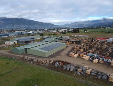 This is a picture of a small industrial area in a rural setting. The foreground is dominated by a large metal shed filled with stacks of wooden pallets. Behind this, there's another building with what appears to be some kind of material storage or production taking place. In the background, you can see a few other buildings and a truck parked outside. There's also a mountain range visible in the distance under a partly cloudy sky. The overall scene suggests an outdoor industrial or warehouse area