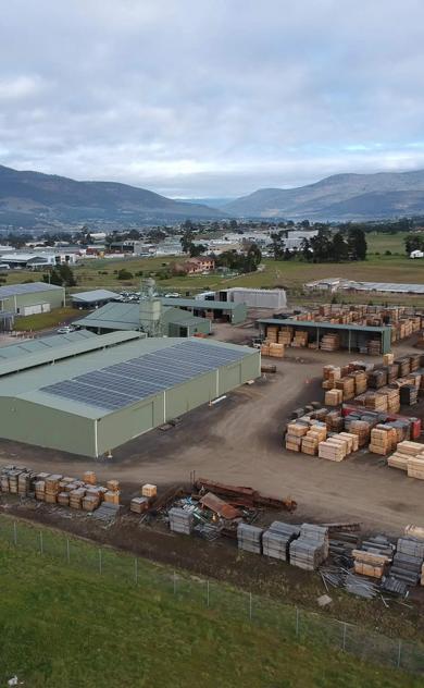 This is a picture of a small industrial area in a rural setting. The foreground is dominated by a large metal shed filled with stacks of wooden pallets. Behind this, there's another building with what appears to be some kind of material storage or production taking place. In the background, you can see a few other buildings and a truck parked outside. There's also a mountain range visible in the distance under a partly cloudy sky. The overall scene suggests an outdoor industrial or warehouse area