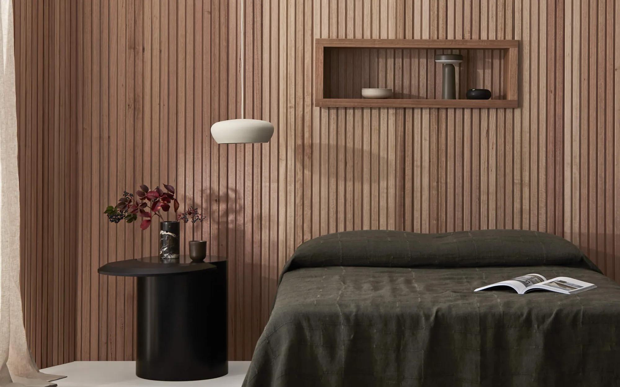 A modern bedroom interior. The room features light-colored Porta wood paneling on the walls and has a natural appearance. There's a dark wooden bed with two pillows, neatly made, positioned in front of a contemporary design nightstand. Above the bed, there are shelves against the wall and a small decorative element on top. A vase with red flowers is visible on the left side of the frame, placed on a surface near the bed. The room has neutral tones, creating an uncluttered and serene atmosphere