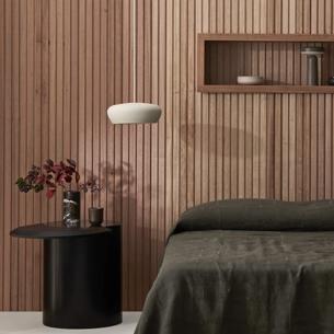 A modern bedroom interior. The room features light-colored Porta wood paneling on the walls and has a natural appearance. There's a dark wooden bed with two pillows, neatly made, positioned in front of a contemporary design nightstand. Above the bed, there are shelves against the wall and a small decorative element on top. A vase with red flowers is visible on the left side of the frame, placed on a surface near the bed. The room has neutral tones, creating an uncluttered and serene atmosphere