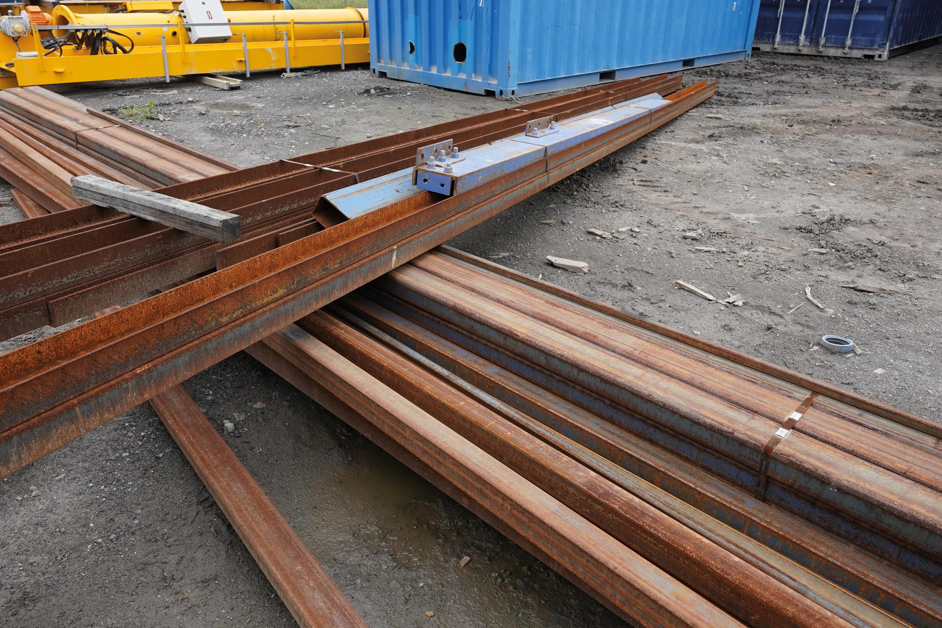 Rusty steel beams and scrap metal at recycling yard with shipping containers.