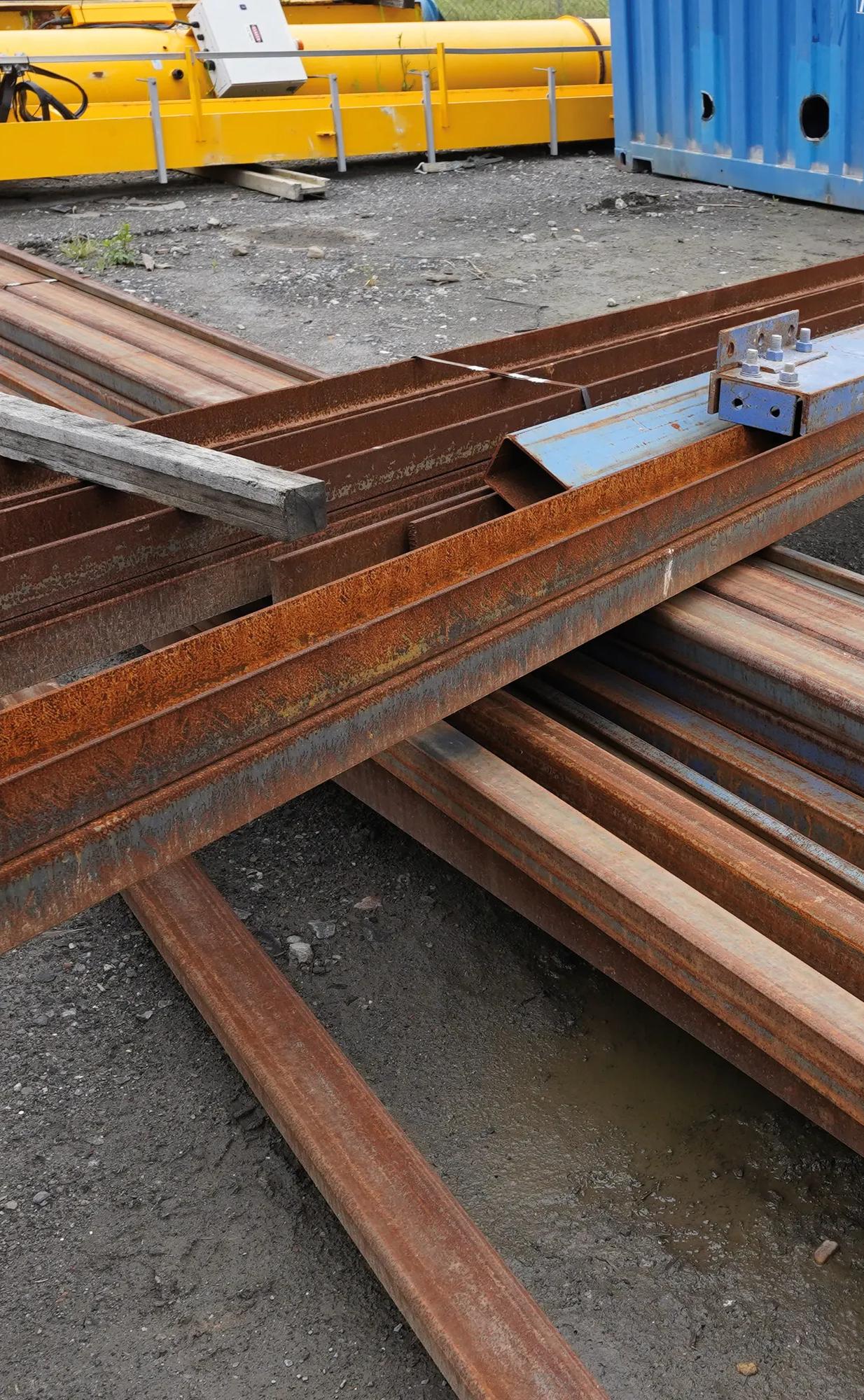 Rusty steel beams and scrap metal at recycling yard with shipping containers.