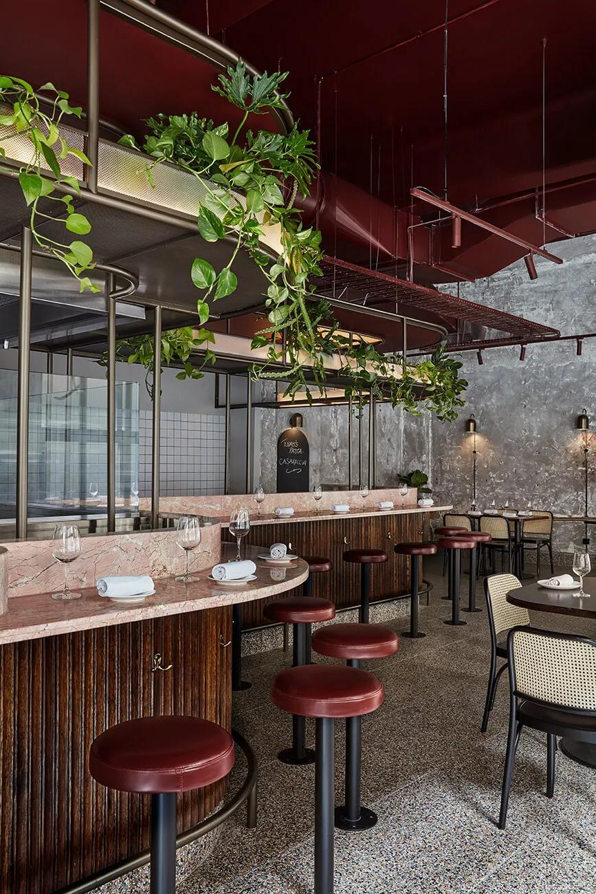 A modern restaurant interior with a contemporary design featuring plants, red ceiling, hanging lights, wooden bar counter, and seating area