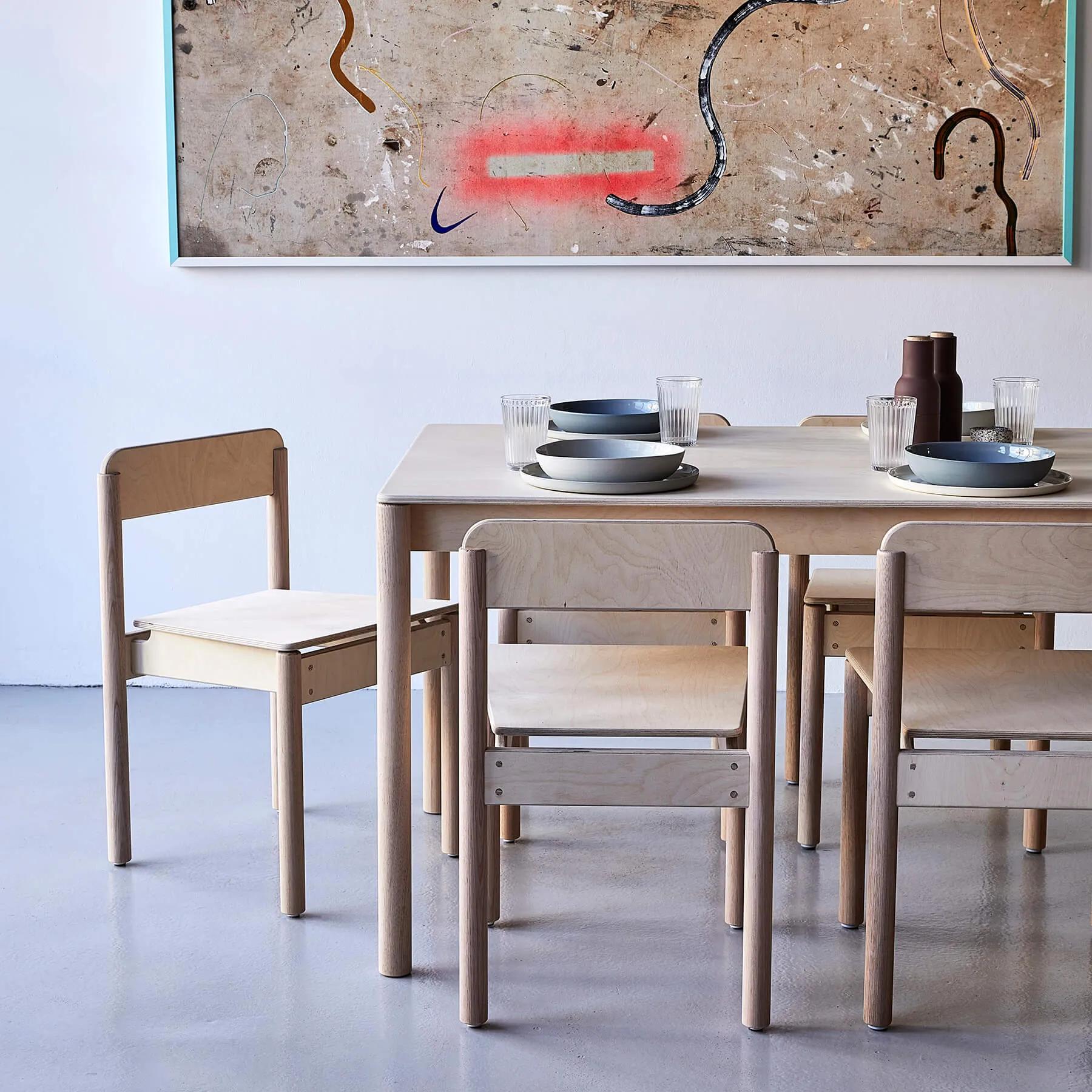 Small wooden dining table set for four, topped with two bowls, plates, and a vase. The table is surrounded by six light-colored chairs