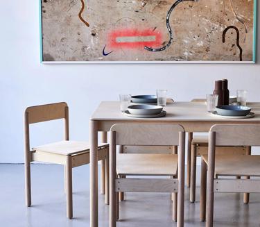 Small wooden dining table set for four, topped with two bowls, plates, and a vase. The table is surrounded by six light-colored chairs