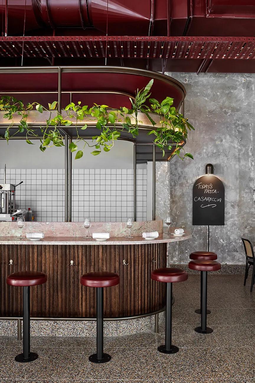 A restaurant interior with a modern design featuring a counter with red stools, black bar stools, a wooden bar, a potted plant, a chalkboard menu, and shelves on the wall