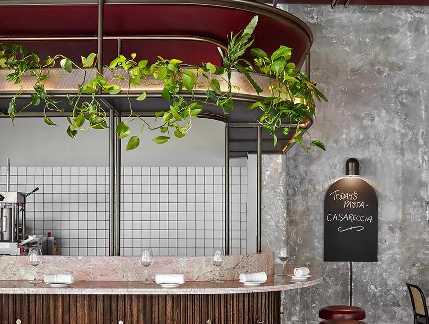A restaurant interior with a modern design featuring a counter with red stools, black bar stools, a wooden bar, a potted plant, a chalkboard menu, and shelves on the wall