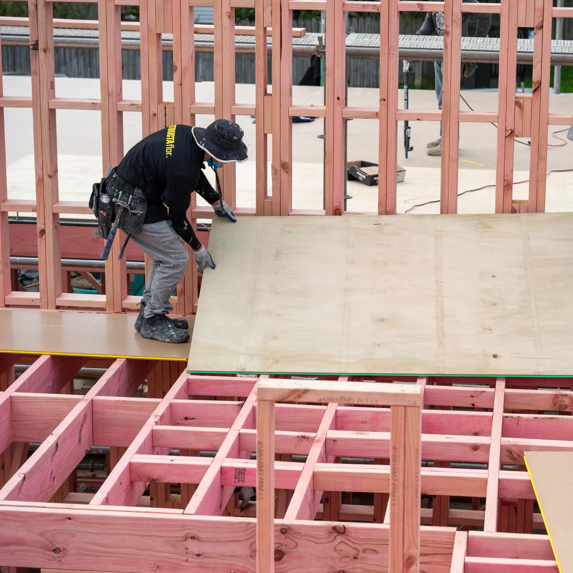 Two workers install a large sheet panel across timber floor framing on a construction site.