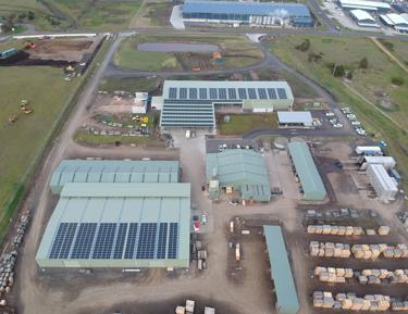 Large building with many windows on ground floor, solar panels attached to roofs of buildings, a large open field in front, multiple trucks parked around perimeter