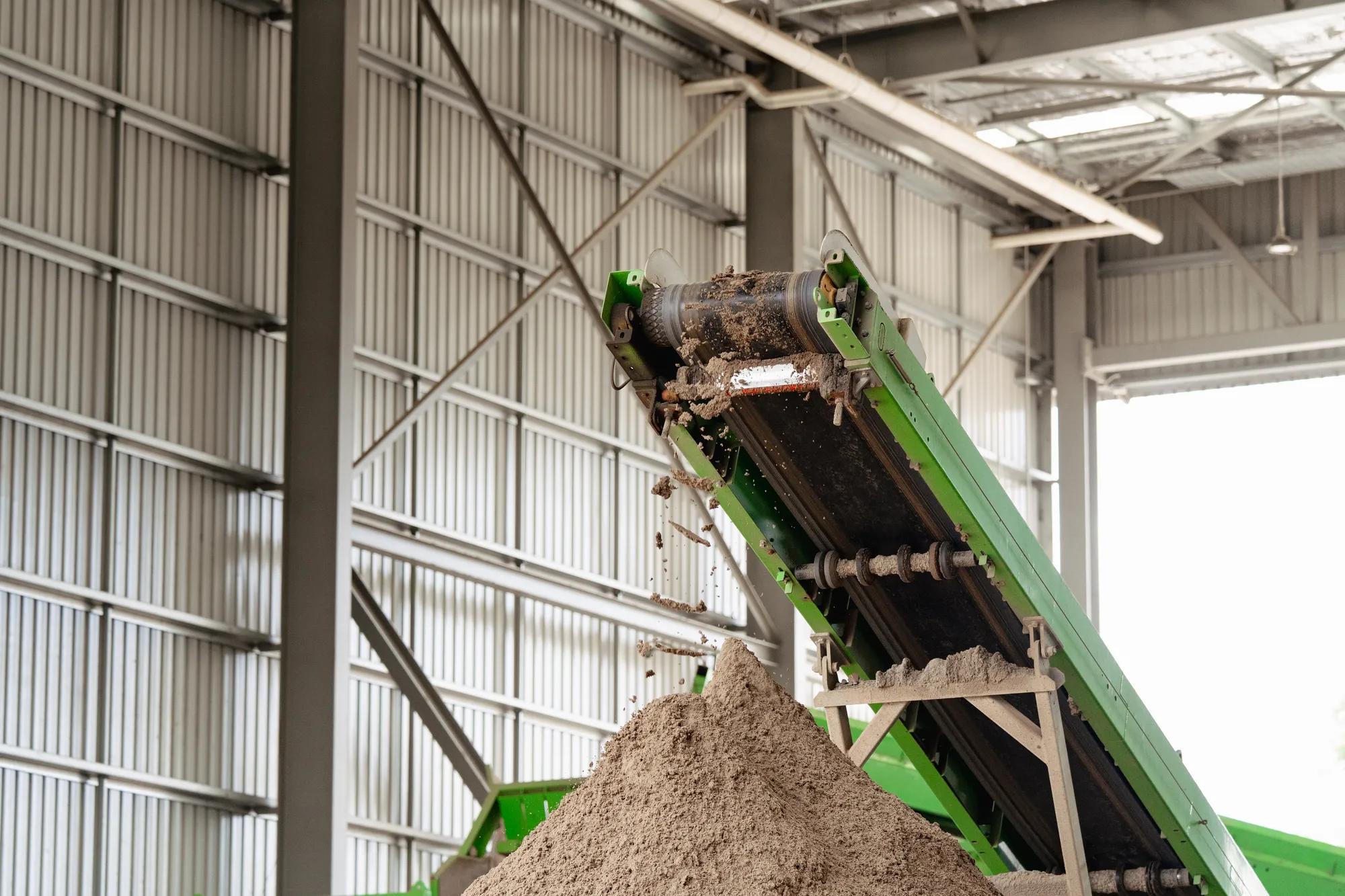 Green conveyor belt depositing processed soil pile inside facility.